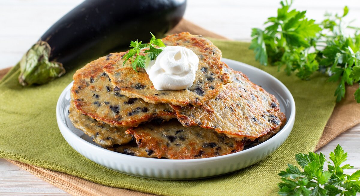 Ein weißer Teller auf einer grünen Tischdecke mit den Auberginen-Pfannkuchen. Rechts vom Bild ein Bund Petersilie und links vom Bild eine Aubergine.