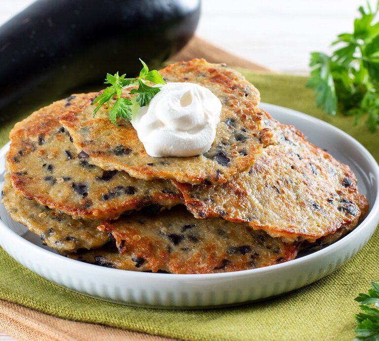 Ein weißer Teller auf einer grünen Tischdecke mit den Auberginen-Pfannkuchen. Rechts vom Bild ein Bund Petersilie und links vom Bild eine Aubergine. | © Shutterstock/ Ahanov Michael