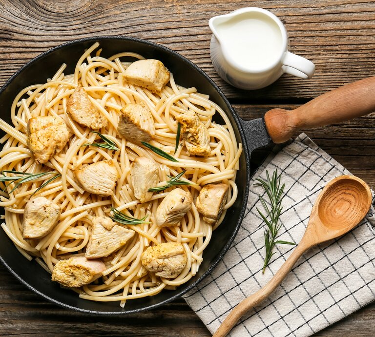 Draufsicht: eine Hähnchen-Nudel-Pfanne mit Rosmarin auf einem Untergrund aus Holz. Daneben liegen ein Tuch und ein Holzlöffel. | © Shutterstock / Pixel-Shot