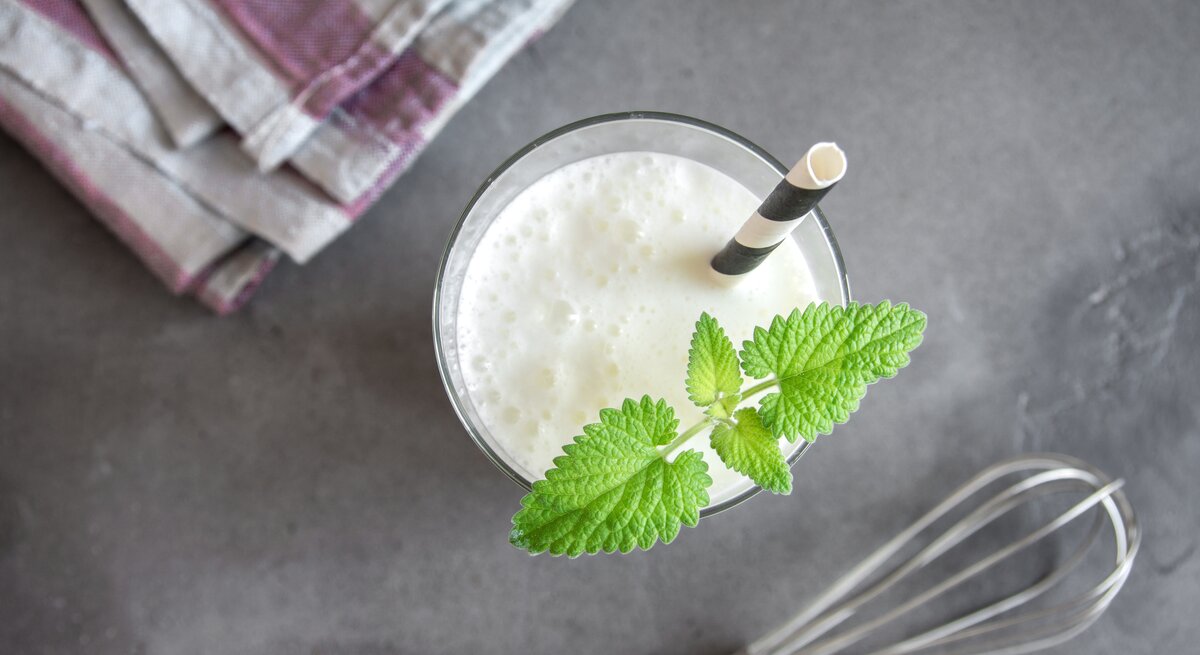  Das Bild zeigt ein Glas Ayran, ein traditionelles türkisches Joghurtgetränk, garniert mit einem frischen Zweig grüner Minze. Es ist von oben aufgenommen, wobei der Fokus auf dem cremigen Getränk liegt. Ein gestreiftes Geschirrtuch und ein Schneebesen liegen auf einer grauen Arbeitsplatte im Hintergrund, was darauf hindeutet, dass das Getränk frisch zubereitet wurde. Ein Papierstrohhalm steckt im Glas, bereit zum Genießen.