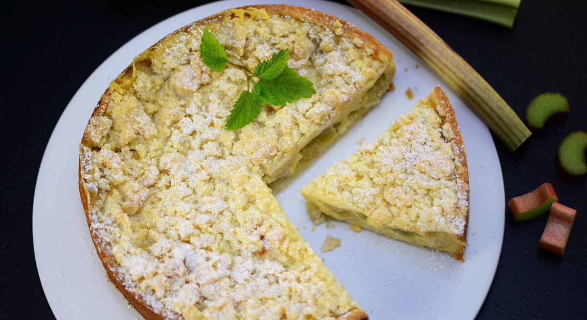 Der Rhabarberkuchen mit einem angeschnittenen Stück und ein paar Rhabarberstangen direkt von oben fotografiert. 