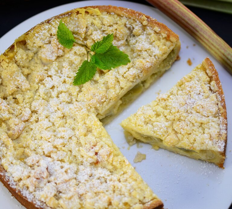 Der Rhabarberkuchen mit einem angeschnittenen Stück und ein paar Rhabarberstangen direkt von oben fotografiert.  | © Shutterstock/ photocrew1