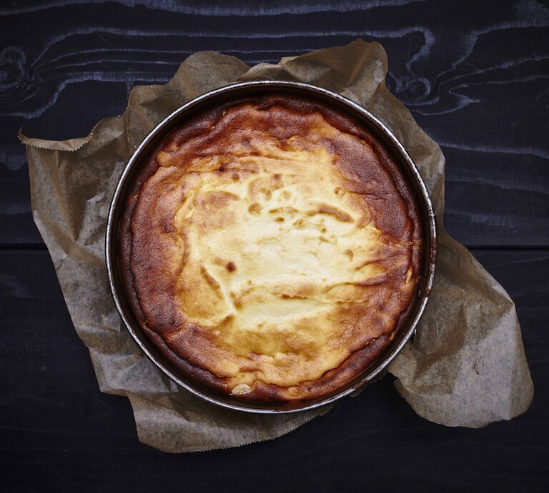 Selbst gebackener Käsekuchen in Form auf einer mit Backpapier ausgelegten, dunklen Arbeitsfläche aus Holz.  | © Getty Images/sorendls