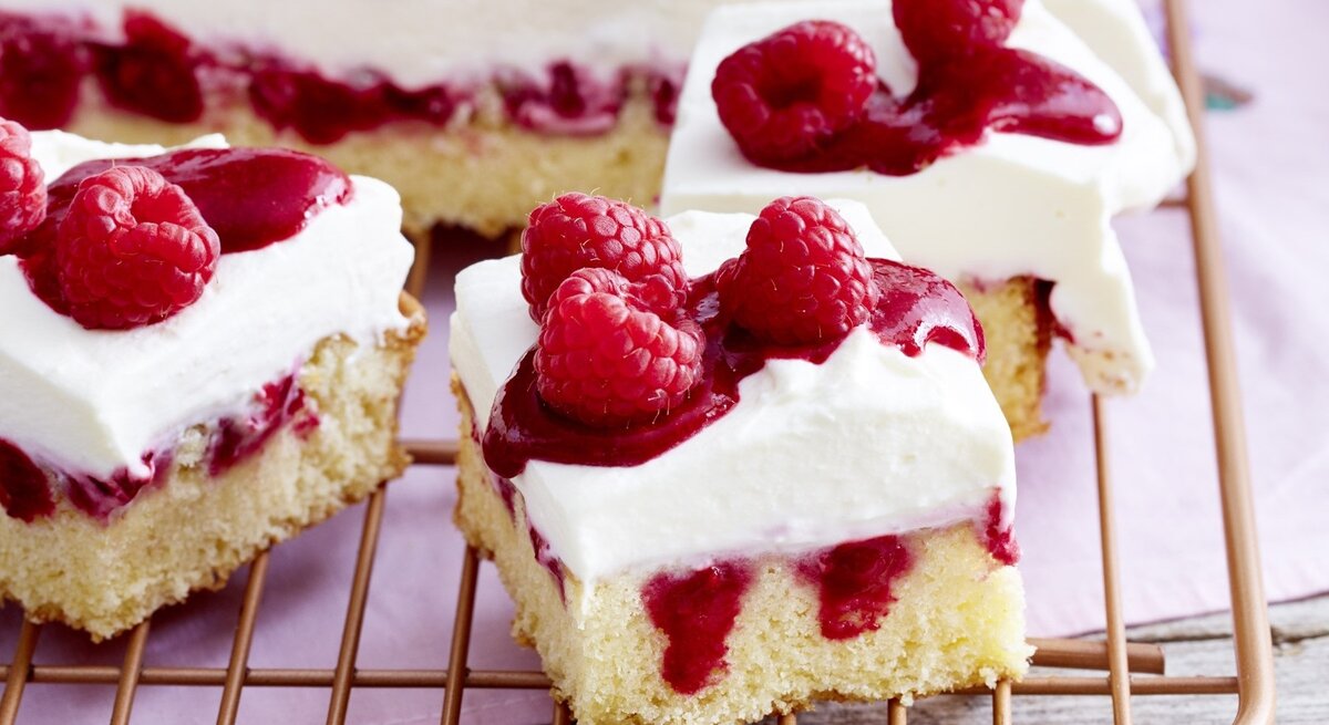 Himbeer-Mascarpone-Kuchen in eckiger Form, angeschnitten auf einem Kuchengitter auf rosanem Tischtuch.