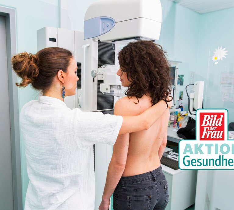 Eine medizinische Fachkraft in weißer Kleidung bereitet eine Mammographie-Untersuchung vor. Die Patientin steht ohne Oberbekleidung an dem Mammographie-Gerät und wird für die Aufnahme positioniert. Die Umgebung wirkt klinisch und professionell, mit einem hellen und sauberen Untersuchungsraum. Auf dem Bild ist auch das Logo von "Bild der Frau AKTION Gesundheit" zu sehen, was die Bedeutung von Vorsorgeuntersuchungen im Gesundheitsbewusstsein unterstreicht. Die Szene vermittelt einen wichtigen Aspekt der Frauengesundheit und präventiven Medizin. | © iStock/choja [M]