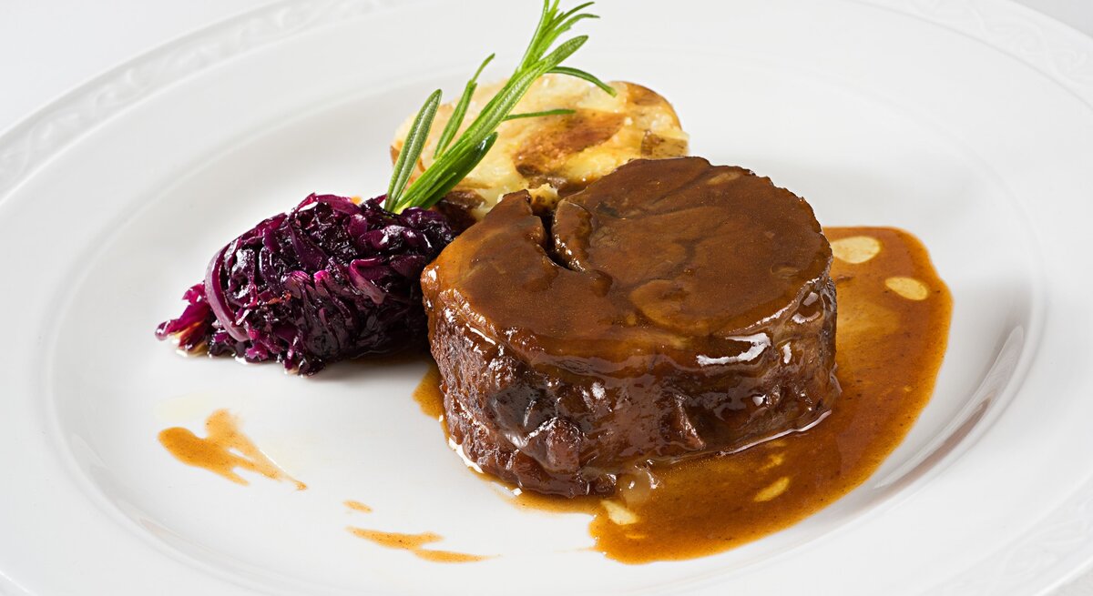 Glühweinbraten mit Soße übergossen, Rotkohl mit Rosmarinzweig und angebratener Knödel auf weißem Teller aus der Draufsicht.