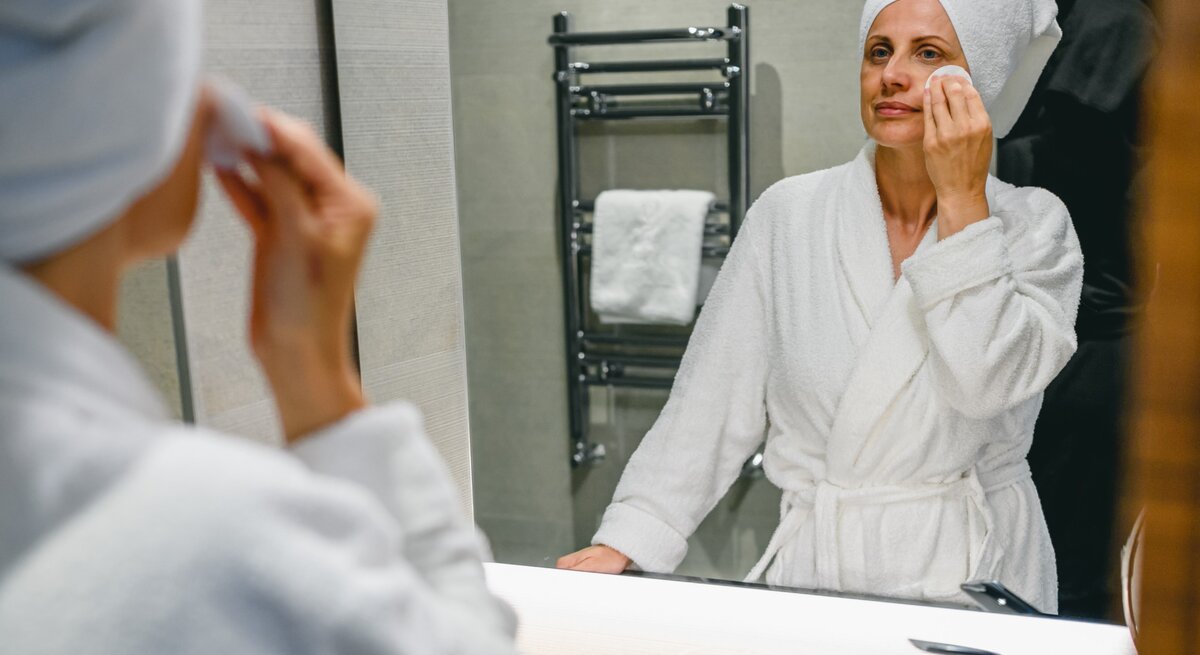 Frau mittleren Alters in einem Badezimmer, die sich selbst im Spiegel betrachtet, während sie ihr Gesicht mit einem Wattepad reinigt. Sie trägt einen weißen Bademantel und ein Handtuch um den Kopf gewickelt, was auf eine Pflegeroutine hindeutet.