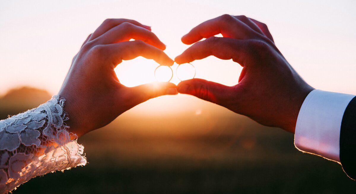 Die Hand einer Frau und die Hand eines Mannes halten Eheringe der untergehenden Sonne entgegen.