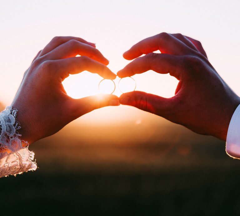 Die Hand einer Frau und die Hand eines Mannes halten Eheringe der untergehenden Sonne entgegen. | © iStock.com/saiva