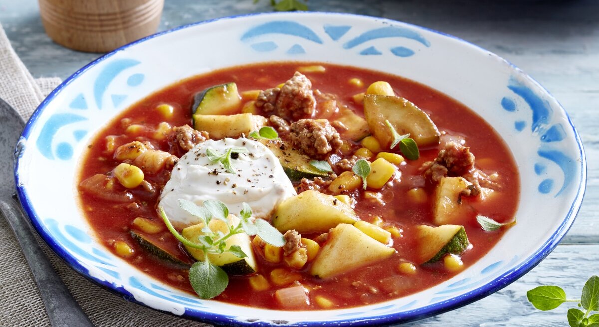 Auf einem tiefen Teller ist eine Portion Zucchini-Tomaten-Eintopf mit Hack serviert. Sie wird mit Frischkäse verfeinert.