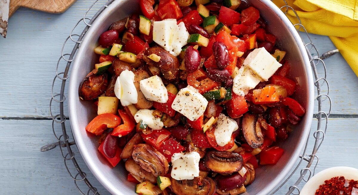 Eine Schüssel Gemüse-Mozzarella-Pfanne in der Draufsicht vor hellem Hintergrund, Chiliflocken, Zwiebel und ein gelbes Küchentuch drum herum. 