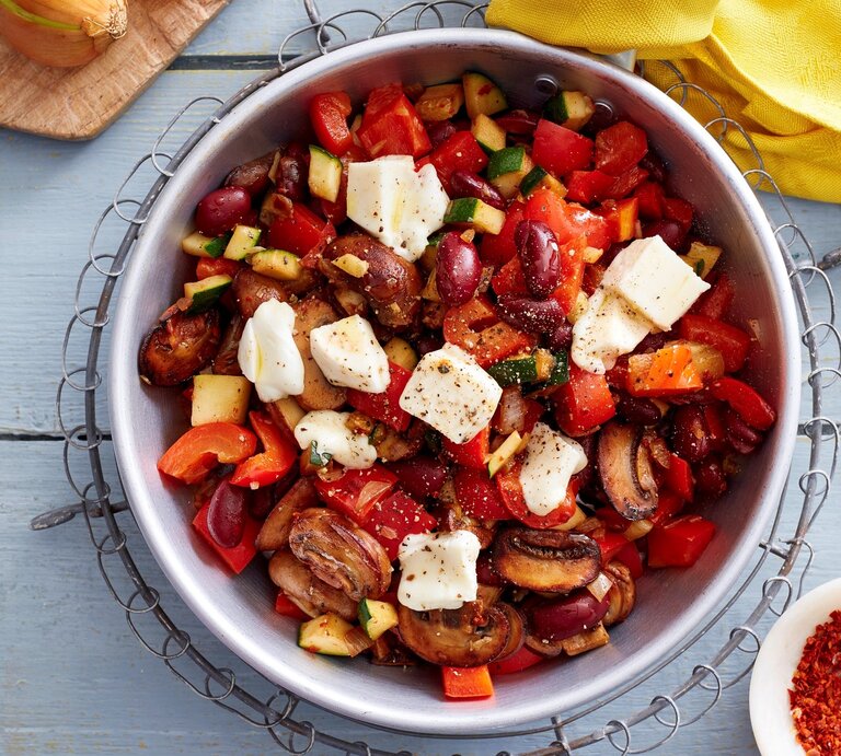Eine Schüssel Gemüse-Mozzarella-Pfanne in der Draufsicht vor hellem Hintergrund, Chiliflocken, Zwiebel und ein gelbes Küchentuch drum herum.  | © EAT CLUB / Isabell Triemer