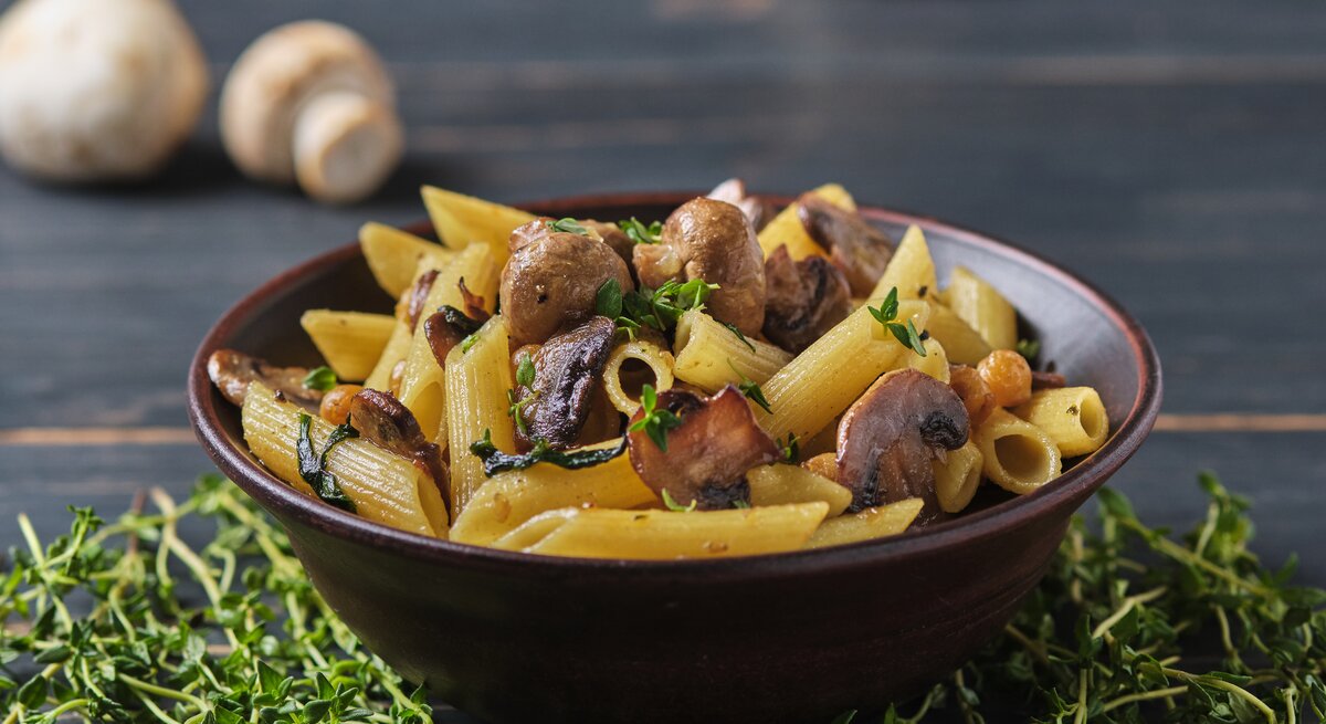 Frontalsicht: Eine dunkle Schüssel mit Penne Rigate mit Thymian-Pilzen steht auf einem dunklen Untergund aus Holz. Daneben liegen frischer Thymian und Champignons.