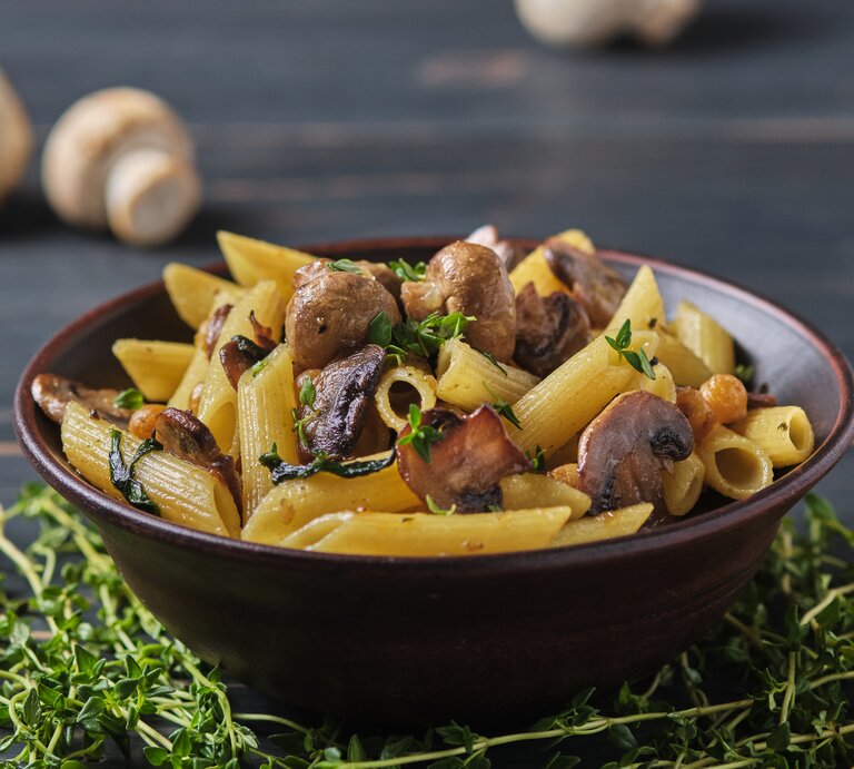 Frontalsicht: Eine dunkle Schüssel mit Penne Rigate mit Thymian-Pilzen steht auf einem dunklen Untergund aus Holz. Daneben liegen frischer Thymian und Champignons. | © Shutterstock / moshko_viktor