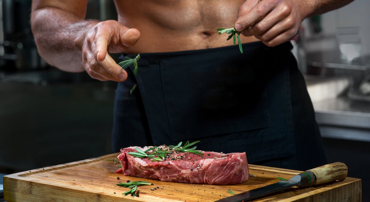 Cedrik Lorenzen: Ein durchtrainierter Mann mit freiem Oberkörper bereitet ein Stück Steak in der Küche zu.