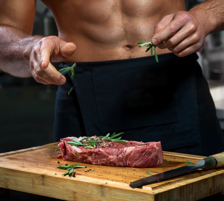 Cedrik Lorenzen: Ein durchtrainierter Mann mit freiem Oberkörper bereitet ein Stück Steak in der Küche zu. | © Shutterstock/ RossHelen