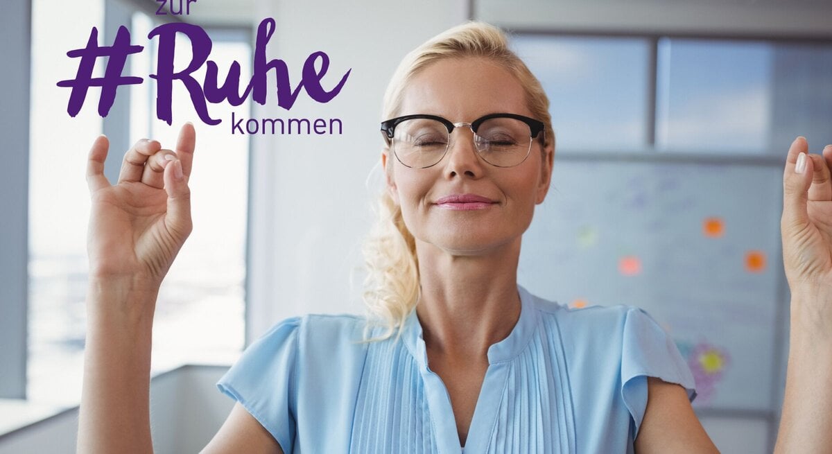 Eine blonde Frau mit geschlossenen Augen und einem zufriedenen Lächeln meditiert in einem hellen Büroumfeld. Sie trägt eine hellblaue Bluse und eine schwarze Brillen. Ihre Hände sind erhoben und formen ein Mudra, eine traditionelle Geste im Yoga, die zur Entspannung beiträgt. Über ihr ist der Text "zur #Ruhe kommen" in lila Schrift zu sehen, was auf Entspannung und Achtsamkeit am Arbeitsplatz hindeutet.