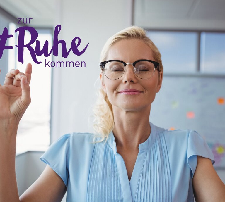Eine blonde Frau mit geschlossenen Augen und einem zufriedenen Lächeln meditiert in einem hellen Büroumfeld. Sie trägt eine hellblaue Bluse und eine schwarze Brillen. Ihre Hände sind erhoben und formen ein Mudra, eine traditionelle Geste im Yoga, die zur Entspannung beiträgt. Über ihr ist der Text "zur #Ruhe kommen" in lila Schrift zu sehen, was auf Entspannung und Achtsamkeit am Arbeitsplatz hindeutet. | © iStock.com/Wavebreakmedia