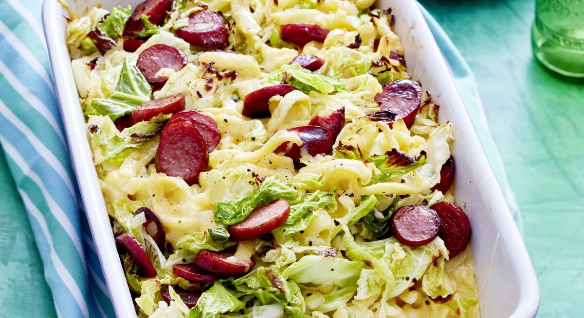 Auflaufform voller Wirsing-Käse-Spätzle mit angeschnittenen Würstchen, serviert auf einem hellen, blauenTisch. 