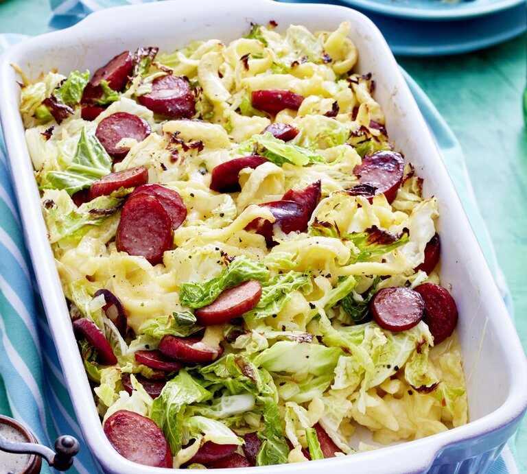 Auflaufform voller Wirsing-Käse-Spätzle mit angeschnittenen Würstchen, serviert auf einem hellen, blauenTisch.  | © EAT CLUB / Isabell Triemer