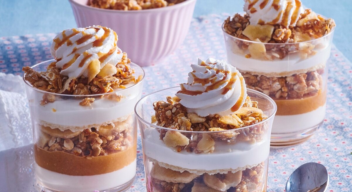 Auf einem Tisch stehen vier Gläser geschichtetes Apfel-Karamell-Parfait. Sie sind mit Sahne und Karamellsoße überzogen. 