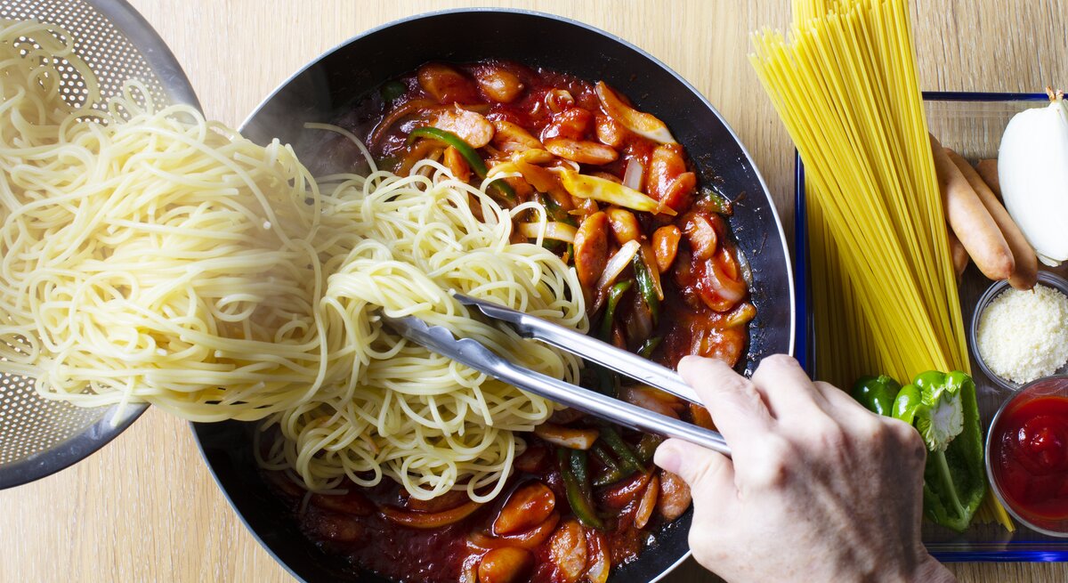 Spaghetti werden aus einem Sieb in eine tomatige Gemüsesoße gekippt.