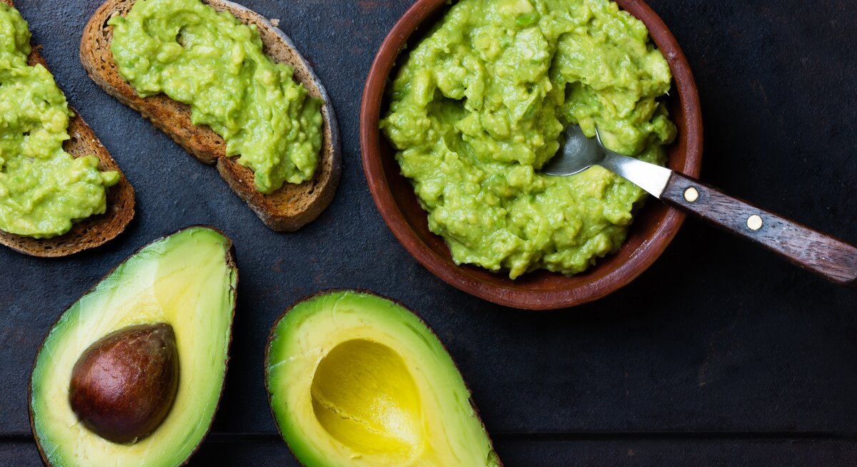 Draufsicht: Zwei Avocado-Hälften liegen auf einem dunklen Hintergrund. Daneben steht eine Guacamole-Soße in einer Tonschüssel. Auch zwei Avocado Sandwiches liegen auf dem Untergrund.