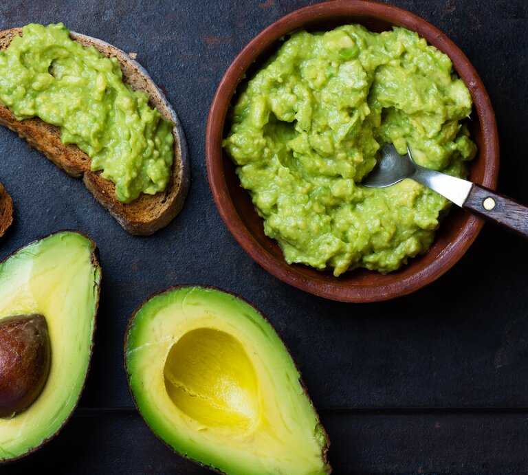 Draufsicht: Zwei Avocado-Hälften liegen auf einem dunklen Hintergrund. Daneben steht eine Guacamole-Soße in einer Tonschüssel. Auch zwei Avocado Sandwiches liegen auf dem Untergrund. | © Shutterstock / Larisa Blinova