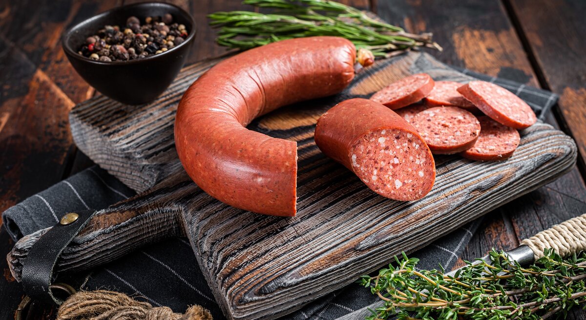Frontal: eine türkische Knoblauchwurst, eine Sucuk, liegt aufgeschnitten auf einem rustikalen Holzbrett. Das liegt auf einem dunklem Holztisch. Drumherum liegen fische Gewürzpfanzen, Gewürze und ein messer.