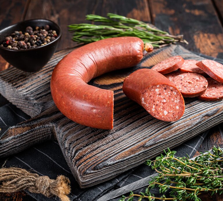 Frontal: eine türkische Knoblauchwurst, eine Sucuk, liegt aufgeschnitten auf einem rustikalen Holzbrett. Das liegt auf einem dunklem Holztisch. Drumherum liegen fische Gewürzpfanzen, Gewürze und ein messer. | © Shutterstock / Mironov Vladimir