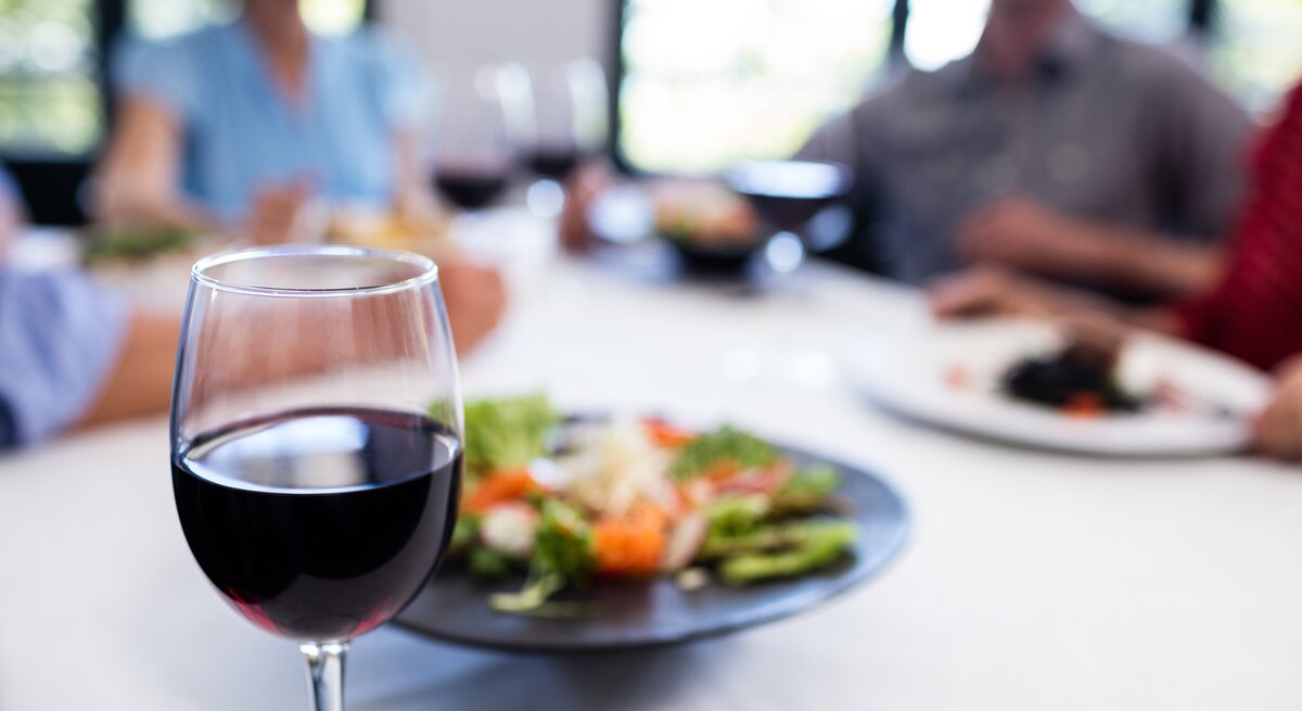 Ein Glas Rotwein im Vordergrund mit scharfem Fokus – im Hintergrund unscharf Personen beim Essen sitzen.