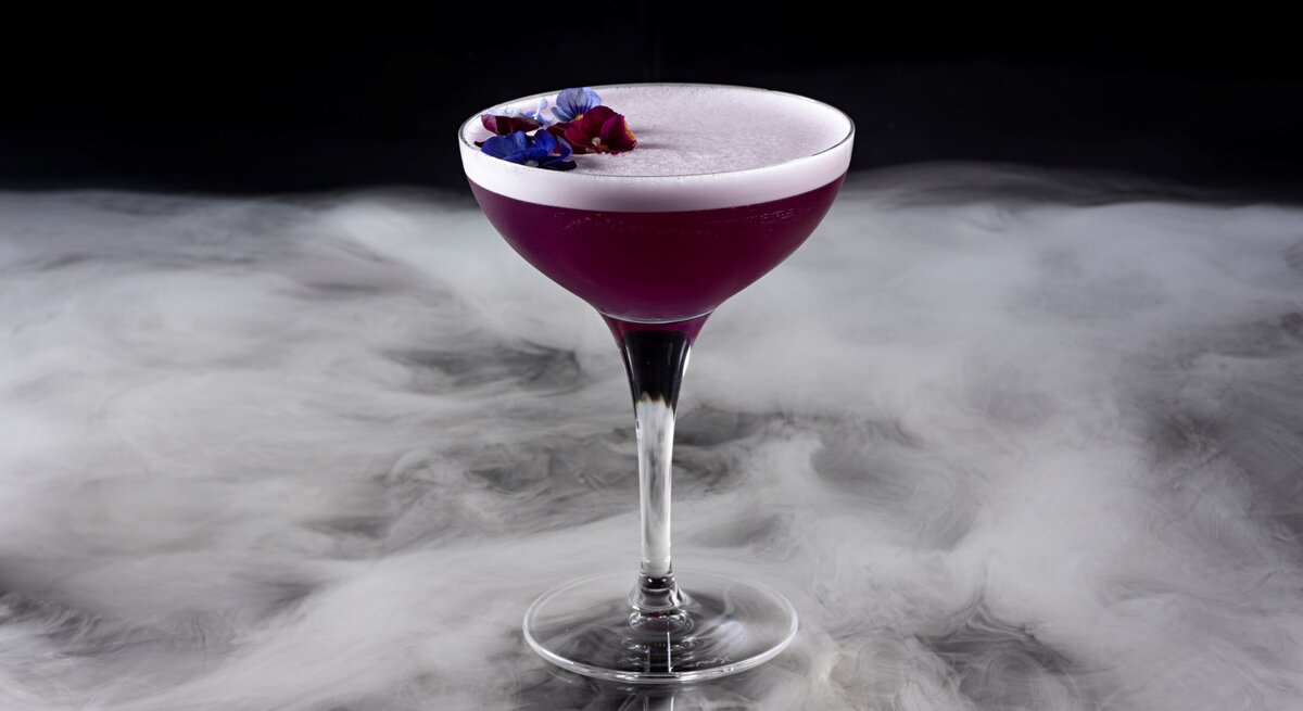 Ein Stielglas mit dem violetten Cocktail auf einem nebelartig-marmoriertem Untergrund.