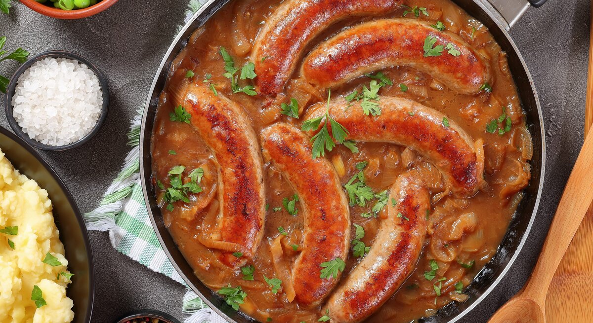 Eine Pfanne mit Bratwürsten in einer braunen Zwiebel-Biersoße. Die Pfanne steht auf einem grün-weiß karierten Geschirrtuch. Daneben ist Salz, Pfeffer, Erbsen und Kartoffelpüree.
