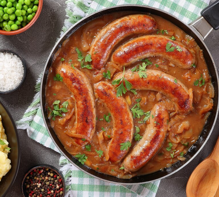 Eine Pfanne mit Bratwürsten in einer braunen Zwiebel-Biersoße. Die Pfanne steht auf einem grün-weiß karierten Geschirrtuch. Daneben ist Salz, Pfeffer, Erbsen und Kartoffelpüree. | © shutterstock/ from my point of view