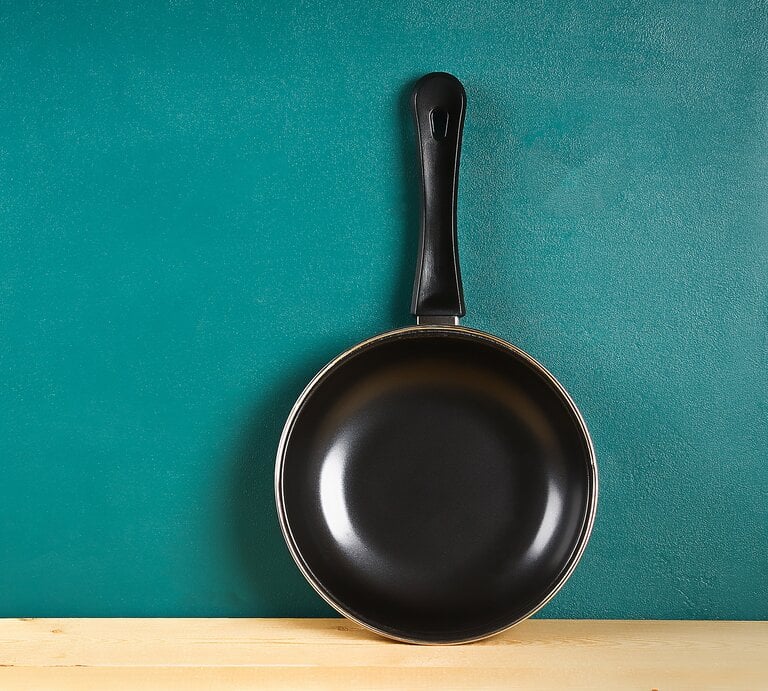 Eine neue schwarze Bratpfanne mit einem schwarzen Kunststoffgriff hängt an einer türkisfarbenen Wand. Die Pfanne ist vorne und im Zentrum positioniert und hängt über einer hellen Holzoberfläche, was eine einfache und saubere Küchenumgebung suggeriert. | © Getty Images/Detry26
