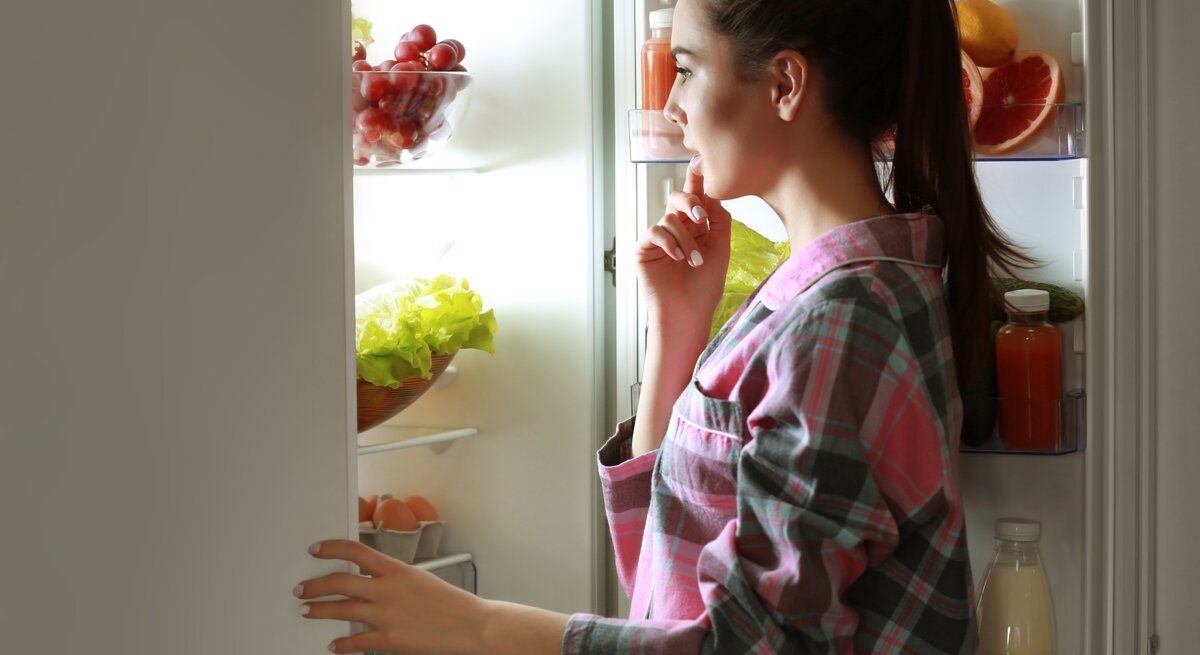 Eine junge, dunkelhaarige Frau, die nachts Hunger spürt, steht an der offenen Kühlschranktür und überlegt, was sie essen kann.