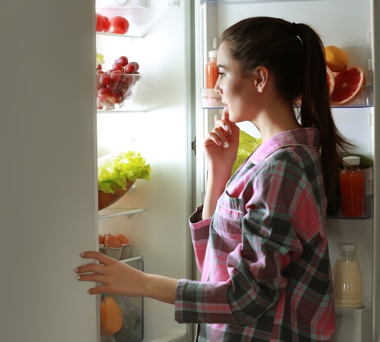 Eine junge, dunkelhaarige Frau, die nachts Hunger spürt, steht an der offenen Kühlschranktür und überlegt, was sie essen kann. | © Shutterstock/ Africa Studio