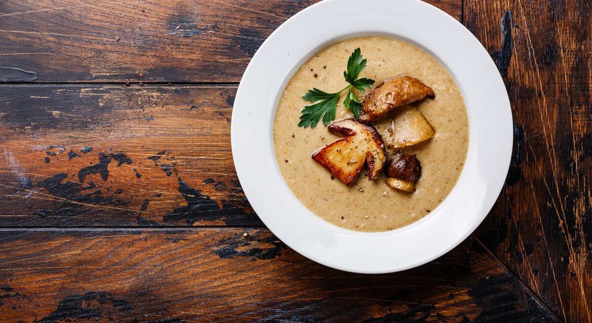 Draufsicht auf einen weißen teller mit cremiger Steinpilzsuppe. Das Ganze steht auf einem dunklen Holztisch. 