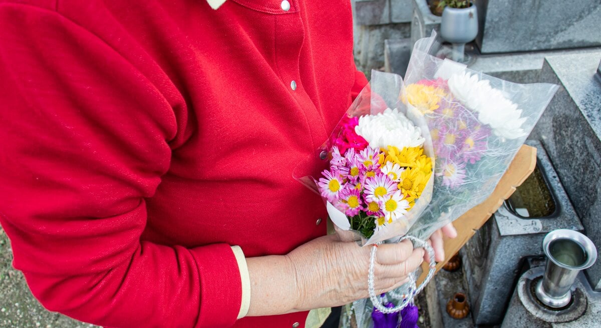 Eine ältere Frau steht mit Blumen in der Hand auf einem Friedhof an einem Grab