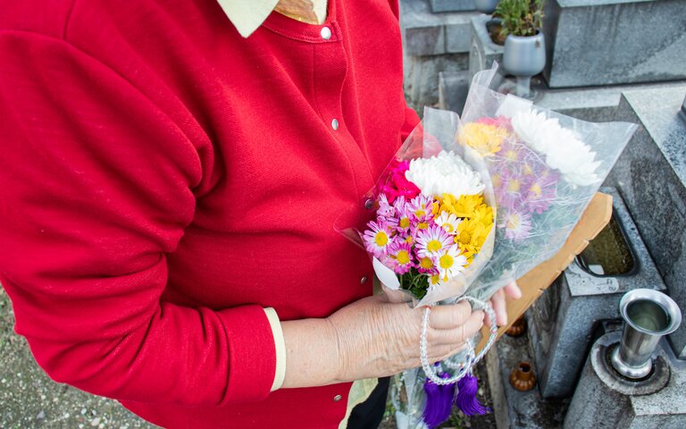 Eine ältere Frau steht mit Blumen in der Hand auf einem Friedhof an einem Grab | © iStock.com/banabana-san