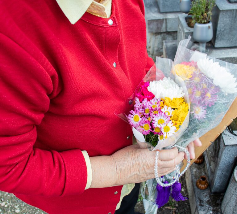Eine ältere Frau steht mit Blumen in der Hand auf einem Friedhof an einem Grab | © iStock.com/banabana-san