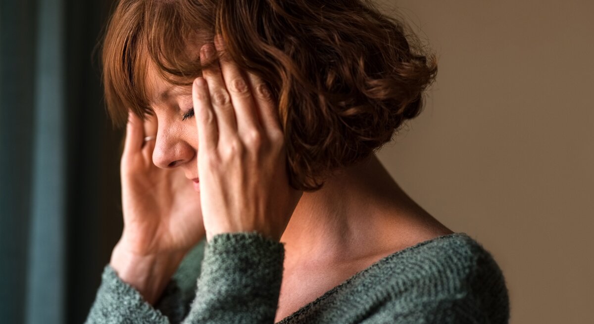  Das Bild zeigt eine Frau mittleren Alters, die sich gestresst oder besorgt zu fühlen scheint. Sie hat ihre Augen geschlossen und ihre Hände an die Schläfen gelegt, was oft eine Geste ist, die Menschen verwenden, wenn sie Kopfschmerzen haben, sich konzentrieren oder versuchen, sich zu beruhigen. Ihr Gesichtsausdruck deutet auf Unbehagen oder Schmerz hin. Sie trägt einen gemütlichen, grünen Pullover, und der Hintergrund ist unscharf, was darauf hinweist, dass der Fokus auf ihrem emotionalen Zustand liegt.