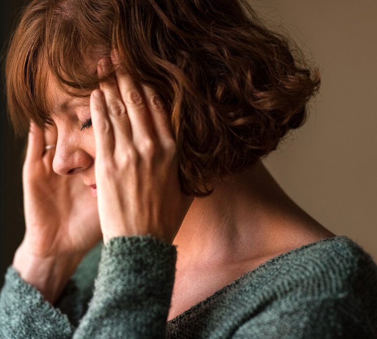  Das Bild zeigt eine Frau mittleren Alters, die sich gestresst oder besorgt zu fühlen scheint. Sie hat ihre Augen geschlossen und ihre Hände an die Schläfen gelegt, was oft eine Geste ist, die Menschen verwenden, wenn sie Kopfschmerzen haben, sich konzentrieren oder versuchen, sich zu beruhigen. Ihr Gesichtsausdruck deutet auf Unbehagen oder Schmerz hin. Sie trägt einen gemütlichen, grünen Pullover, und der Hintergrund ist unscharf, was darauf hinweist, dass der Fokus auf ihrem emotionalen Zustand liegt. | © iStock.com/NickyLloyd