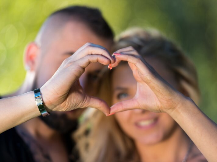Pärchen, das mit ihren Händen ein Herz formt und durch dieses blickt, vor einem verschwommenen, grünen Hintergrund. | © iStock.com/Ibrakovic