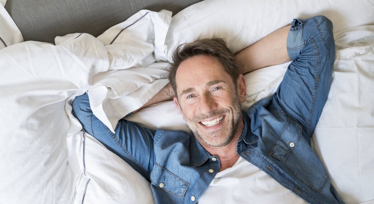 Ein Mann hat die Hände hinter dem Kopf verschränkt und liegt entspannt auf einem Bett. Er trägt ein Jeans-Hemd und lächelt.