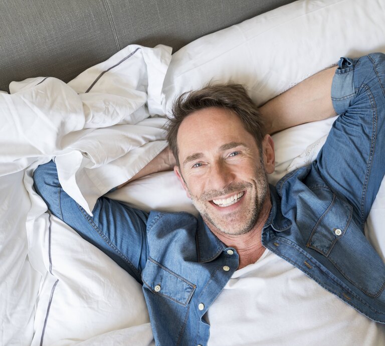 Ein Mann hat die Hände hinter dem Kopf verschränkt und liegt entspannt auf einem Bett. Er trägt ein Jeans-Hemd und lächelt. | © imago/Westend61