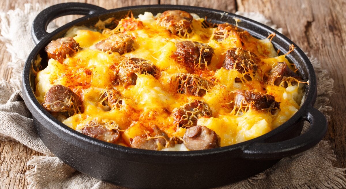 Draufsicht, schräg: auf Holzdielen steht eine runde und dunkle Auflaufform mit Griffen. Das Gericht heißt Jolean's Cheese Potato, es handelt sich um ein amerikanischen Auflauf mit Cabanissi-Würstchen.