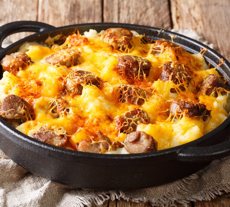 Draufsicht, schräg: auf Holzdielen steht eine runde und dunkle Auflaufform mit Griffen. Das Gericht heißt Jolean's Cheese Potato, es handelt sich um ein amerikanischen Auflauf mit Cabanissi-Würstchen. | © Shutterstock / AS Foodstudio