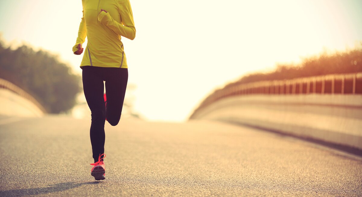 Ein Läufer in auffälliger Sportbekleidung joggt auf einer Straße, umgeben von sanftem Sonnenlicht und einer ruhigen Landschaft, was auf Training bei Tagesanbruch oder Sonnenuntergang hindeutet.