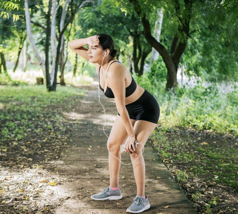 Eine Frau in Sportbekleidung macht eine Pause beim Joggen in einem grünen Park. Sie hält sich die Hand an die Stirn, hat Kopfhörer in den Ohren und sieht erschöpft aus. | © IMAGO / imagebroker